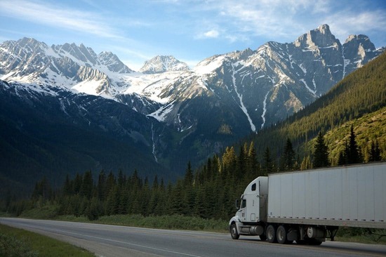 White semi-truck driving along a scenic highway with a backdrop of snow-capped mountains and dense forest.