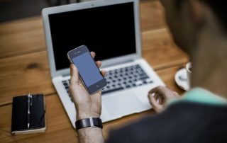 Person holding a smartphone while working on a laptop at a wooden desk, with a notebook and pen placed beside.