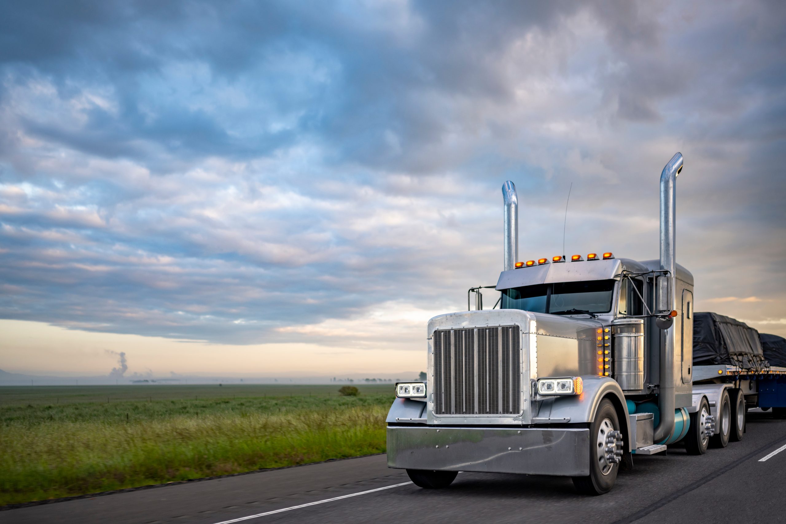 Silver semi-truck hauling cargo on a highway through a rural landscape under a cloudy sky, with fields stretching into the distance.