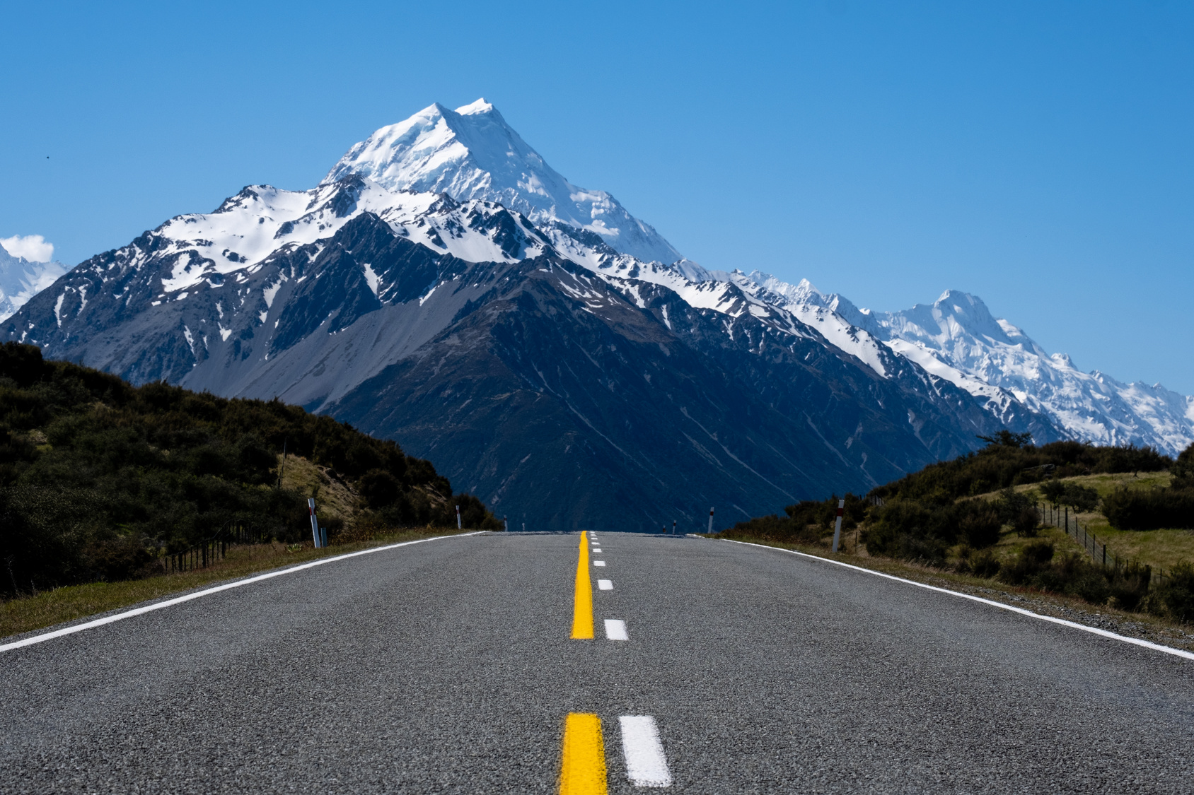 Open road leading to a snow-covered mountain range under a clear blue sky.