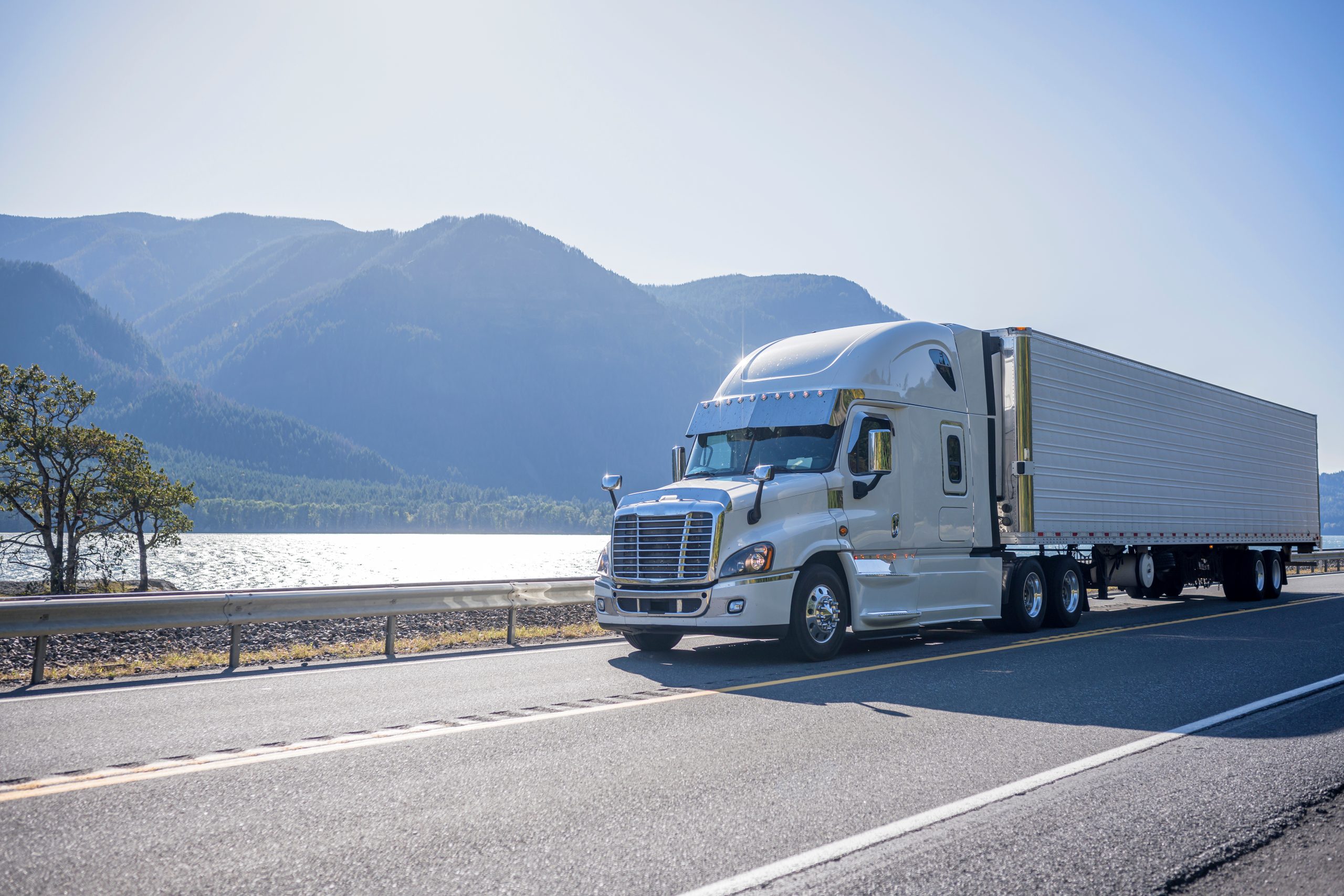 Semi-truck driving on a scenic road by a lake and mountains.