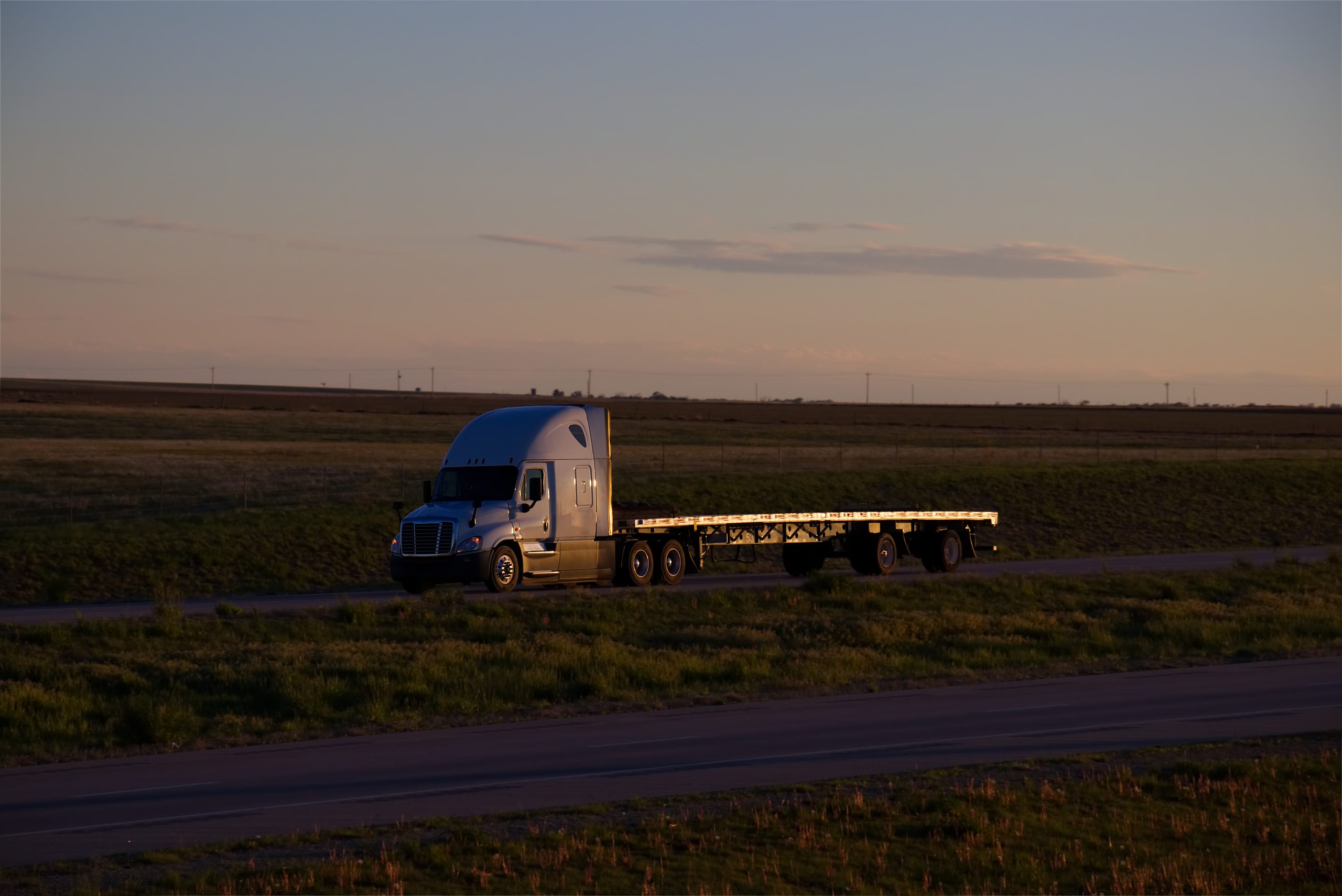 Blue semi-truck with an empty flatbed trailer driving through an open field at sunset.