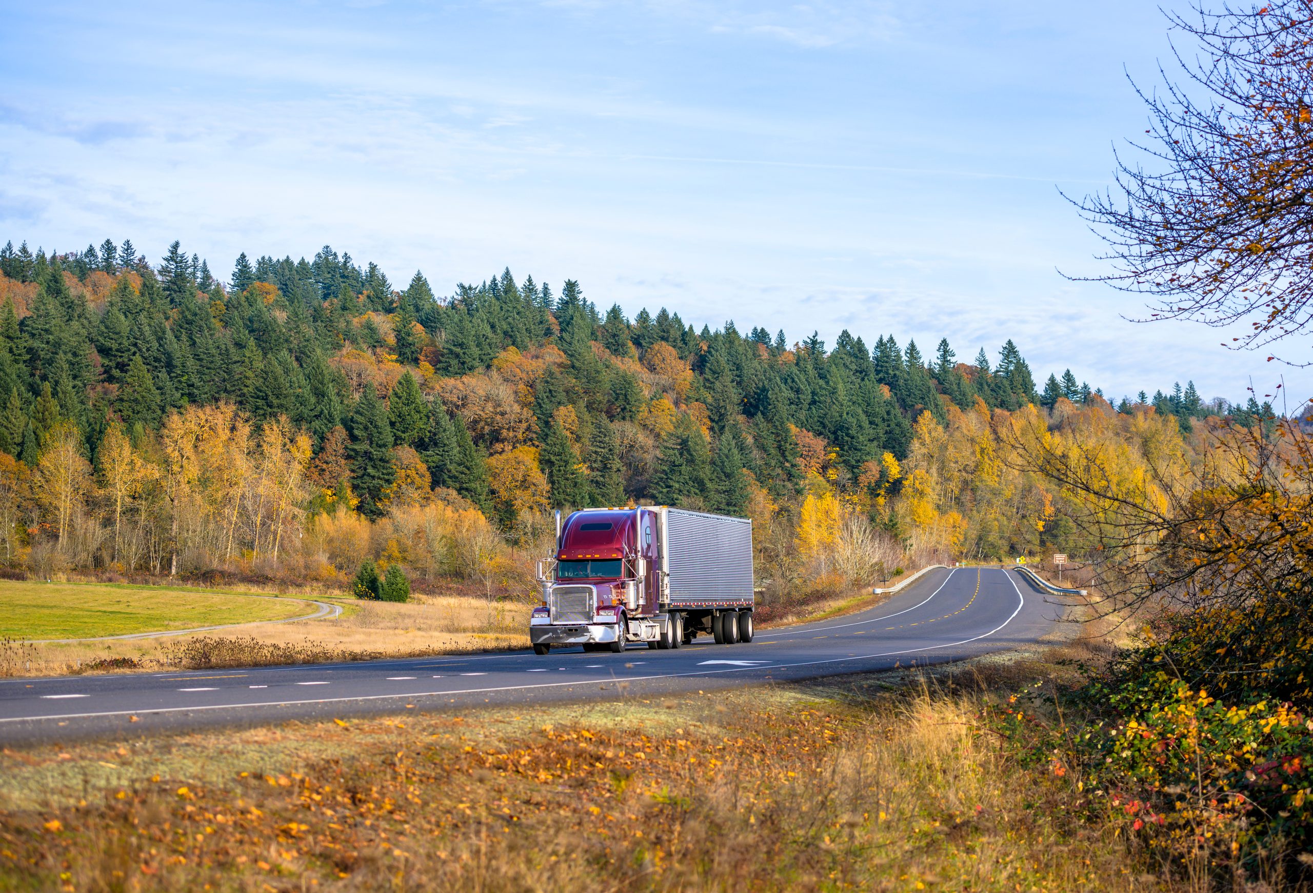 Red semi-truck driving along a scenic rural road surrounded by colorful autumn trees.