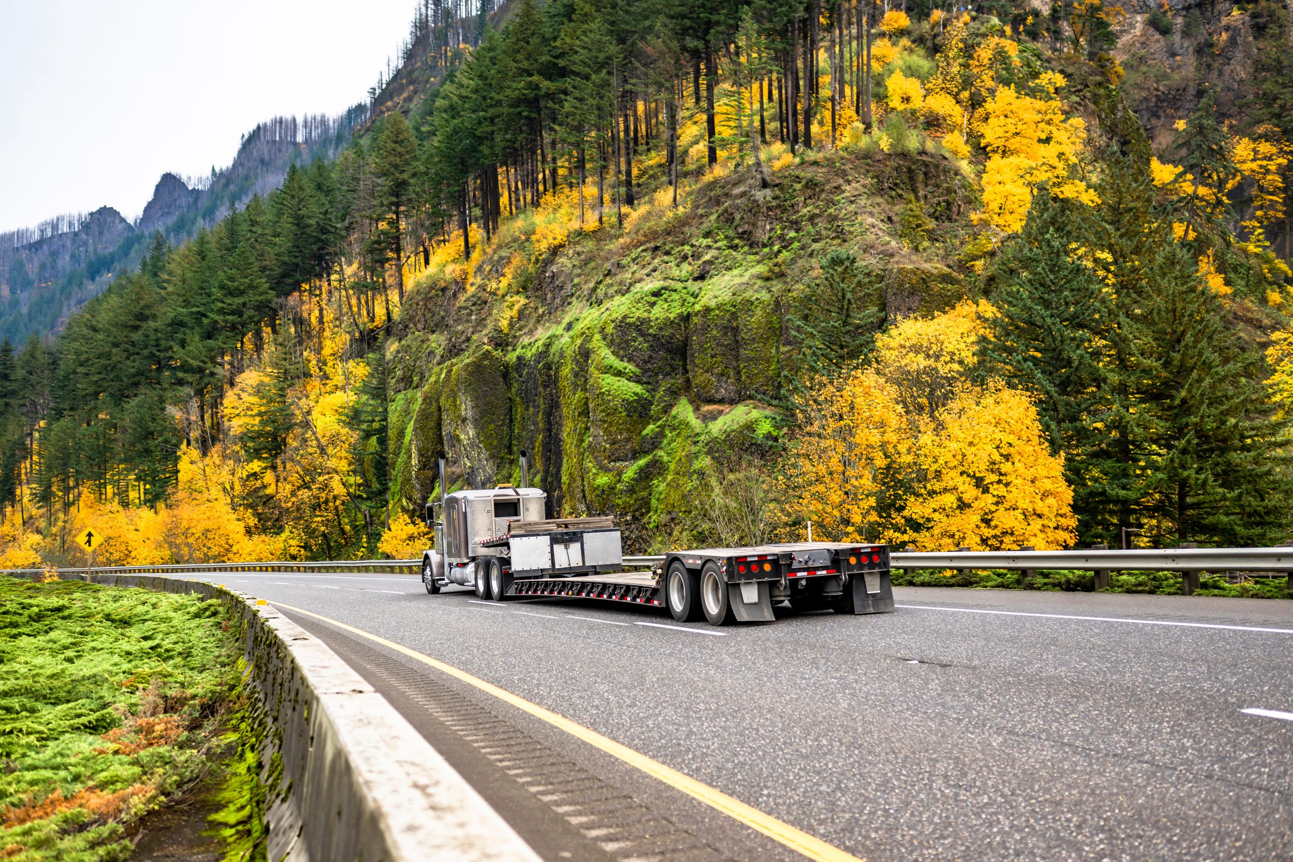 Silver semi-truck with an empty flatbed trailer driving along a mountain road with vibrant yellow and green autumn foliage.