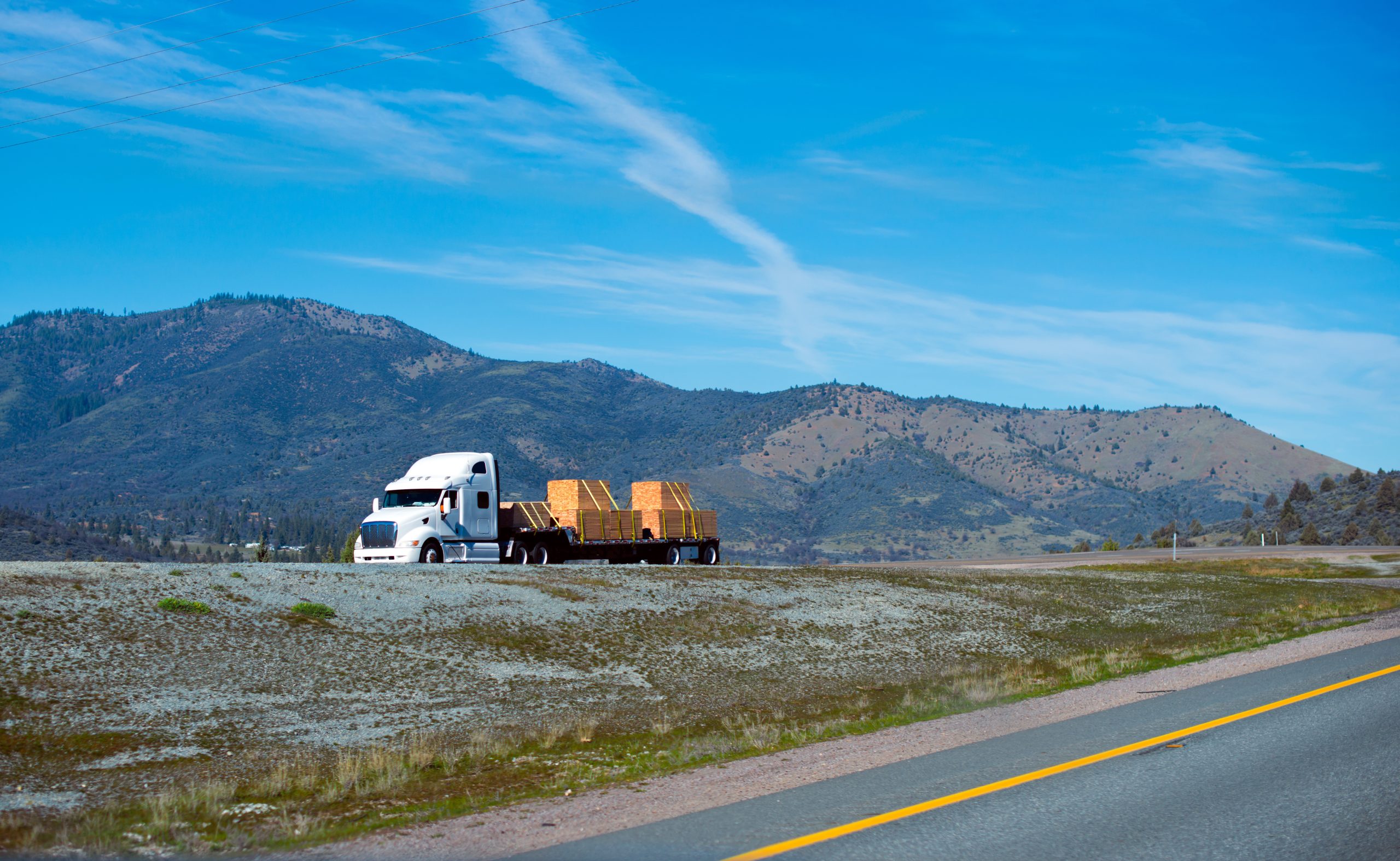 White semi-truck hauling a flatbed trailer loaded with wooden pallets on a highway, set against a backdrop of mountainous terrain under a clear blue sky.