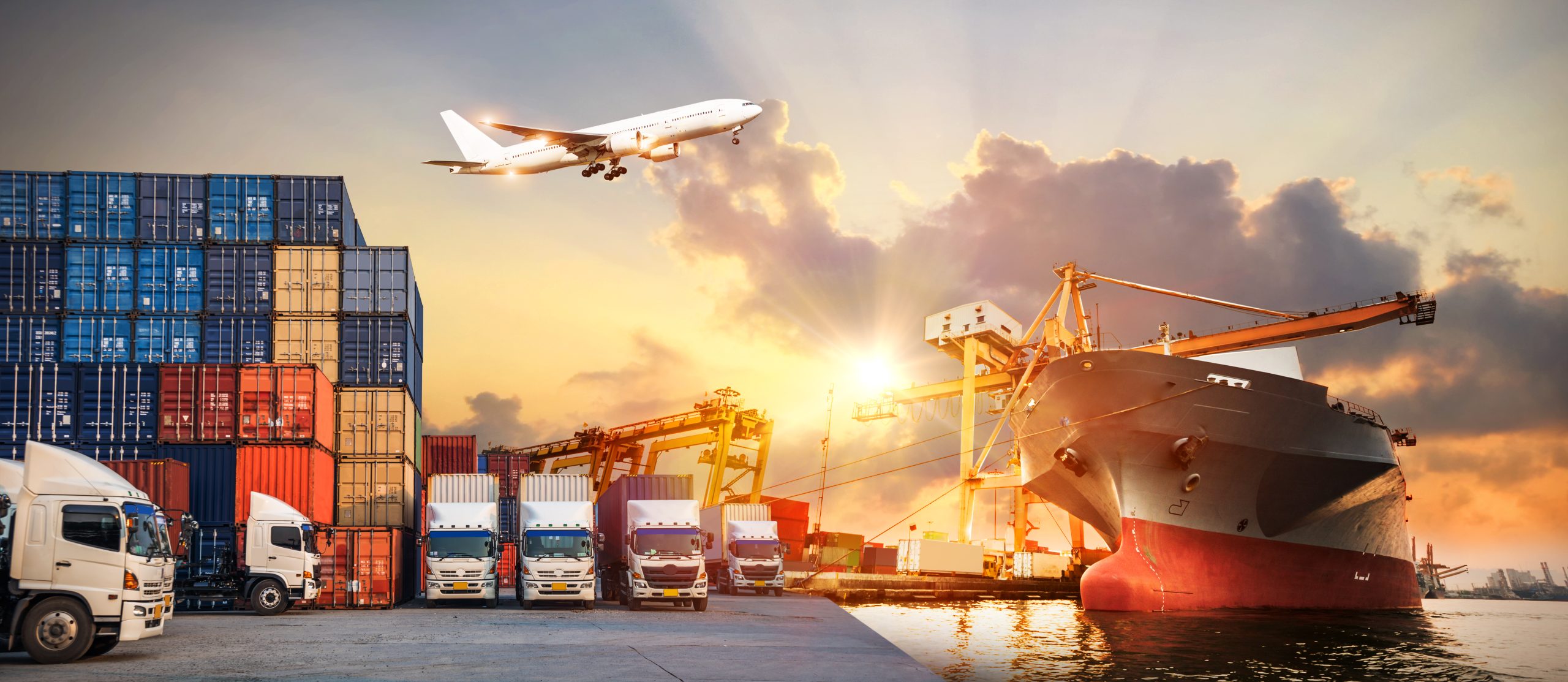 Multimodal logistics hub with trucks, stacked cargo containers, a large shipping vessel, and an airplane taking off in the background at sunset.